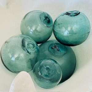 May include: A collection of four spherical glass fishing floats in varying sizes, featuring a translucent teal-green color. Each orb displays a distinct circular seal or pontil mark on its surface, characteristic of hand-blown glass construction.