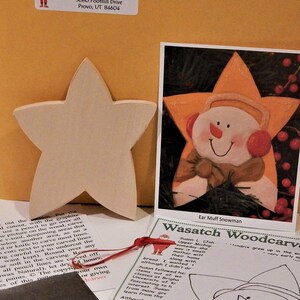 May include: A wooden star with a snowman painted on it. The snowman is wearing a red scarf and a brown hat. The star is painted yellow and has a red border. The text "Ear Muff Snowman" is written below the image. The text "Wasatch Woodcarver" is written on a separate card with a snowman illustration.