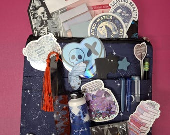 May include: A blue and black zippered pouch with a variety of stickers, pins, and other small items. The pouch has a starry pattern and the text "Pretty Girls Road" is visible on one of the stickers. The text "Leaving Reality" is visible on one of the pins.