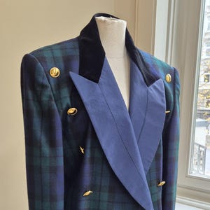 May include: A double-breasted blazer in a green and blue plaid pattern. The blazer features a navy blue lapel and collar, with gold-colored buttons. The jacket is displayed on a mannequin.