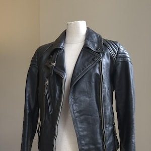May include: Black leather jacket with an asymmetrical zipper closure, lapel collar, and quilted shoulder detailing. The jacket is displayed on a mannequin, showcasing its design and fit. A classic biker style.