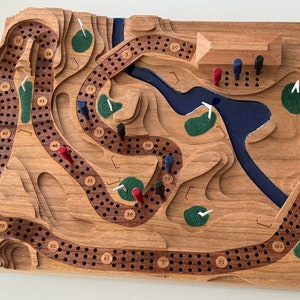 Peut inclure: Un plateau de jeu en bois avec un design de terrain de golf. Le plateau présente un lac bleu, des terrains de golf verts et des drapeaux de golf blancs. Le plateau est en bois et a une finition naturelle. Le plateau est numéroté de 1 à 115. Il y a des pions rouges et bleus sur le plateau.