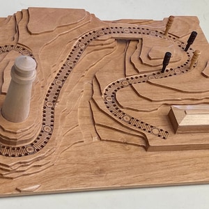 May include: A wooden game board with a lighthouse, a house, and a winding path. The path is lined with small circles and the board is made of light wood.