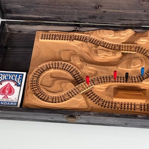 Könnte beinhalten: Ein Holzspielbrett mit einem geschnitzten Bahngleisdesign. Das Brett befindet sich in einer Holzkiste mit schwarzer Oberfläche. Auf dem Gleis befinden sich rote und blaue Spielfiguren.