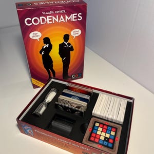 May include: The box for the Codenames word game is open, revealing the game components. The box is red and features a silhouette of two people. The game components include a timer, a grid of colored squares, and a stack of cards.