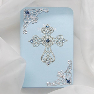 Puede incluir: Una tarjeta azul claro con una cruz plateada y adornos en las esquinas. La cruz y las esquinas están adornadas con piedras preciosas azules. La palabra "Testamento" es visible en la tarjeta.