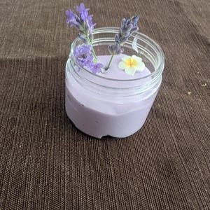 Puede incluir: Un pequeño tarro de cristal transparente lleno de crema de color púrpura claro. Ramas de lavanda y una pequeña flor blanca y amarilla están dispuestas encima de la crema. El tarro está sobre una superficie marrón texturizada.