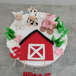 Könnte beinhalten: Ein roter und weißer Scheunen-Kuchentopper mit Kuh-, Schwein- und Schaf-Figuren. Der Kuchen ist mit grünem Gras, weißen Blumen und einer rosa Blume verziert. Die Zahl 3 und der Name "MIRAC" sind ebenfalls auf dem Kuchen.