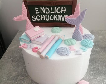 May include: A white cake decorated with a chalkboard sign that says "ENDLICH SCHULKINI", mermaid tails, books, pencils, and seashells.