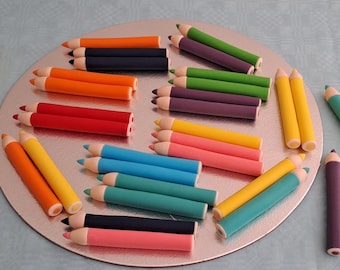 May include: A collection of colorful pencils arranged on a white plate. The pencils are made of fondant and are decorated with different colors.