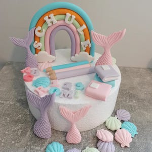 Könnte beinhalten: Ein weißer Kuchen, der mit einem Regenbogen, Meerjungfrauen-Schwänzen, Muscheln, Bleistiften und einem Buch verziert ist. Der Regenbogen hat das Wort "SCHULE" darauf geschrieben.