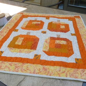 May include: A small orange and white patchwork quilt with a design of four pumpkins. The quilt is made with a variety of fabrics, including orange, white, and yellow.