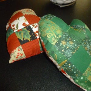 May include: Two patchwork heart-shaped pillows. One pillow is red, green, and white with a holly leaf pattern. The other pillow is green with a gold and white snowflake pattern.