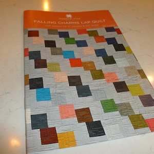May include: A brochure titled "Falling Charms Lap Quilt" features a quilt pattern with a gray background and colorful square blocks. The squares are in shades of red, blue, green, orange, and brown. The brochure has an orange top.