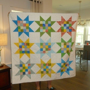 May include: A white quilt with nine colorful star blocks. Each star block is made of a patchwork of squares in shades of blue, green, yellow, and red. The quilt is being held up by a person.