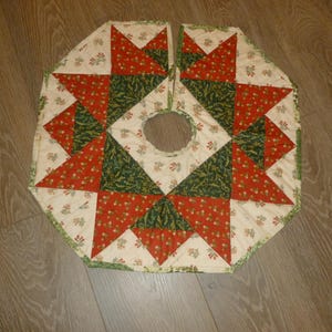 May include: A quilted Christmas tree skirt with a starburst design. The skirt features red and green triangles, floral accents, and a cream-colored background. The skirt has a diameter of approximately 40 inches.