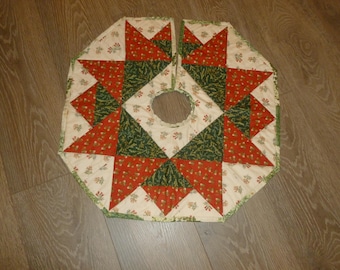 May include: A quilted Christmas tree skirt with a starburst design. The skirt features red and green triangles, floral accents, and a cream-colored background. The skirt has a diameter of approximately 40 inches.