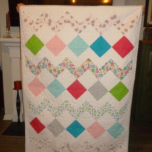 May include: A white quilt with a diamond pattern in various colors, including pink, green, blue, and gray. The quilt has a zigzag pattern with floral and patterned fabric accents. The edges are finished with a decorative trim.