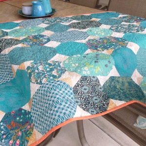 May include: A handmade quilt featuring a patchwork design of teal, turquoise, and patterned fabrics. The quilt has an orange border and is displayed on a table. The quilt's design includes floral and geometric patterns.