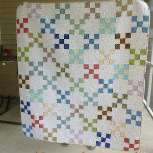 May include: A square quilt featuring a white background with a repeating pattern of small, multi-colored fabric squares arranged in cross shapes. The quilt displays various colors including red, blue, green, brown, yellow, and light purple.