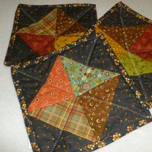 May include: Two square quilted potholders featuring a dark charcoal background with a yellow berry print. Each potholder displays a central star pattern composed of various fabric triangles in shades of orange, green, brown, and plaid.