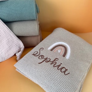 Könnte beinhalten: Eine hellbeige Strickdecke mit dem Namen "Sophia" in brauner Schrift gestickt, zusammen mit einem Regenbogen-Design in Braun, Rosa und Weiß. Im Hintergrund sind mehrere andere gefaltete Decken in Grün- und Beigetönen gestapelt.