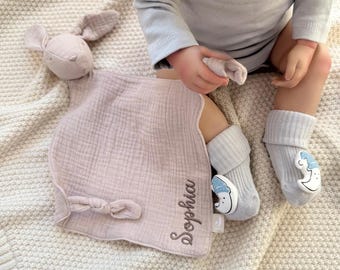 May include: A light purple baby security blanket with a bunny head and a bow. The blanket is personalized with the name "Sophia" embroidered in a matching thread. Gray socks with a moon and cloud design complete the set.