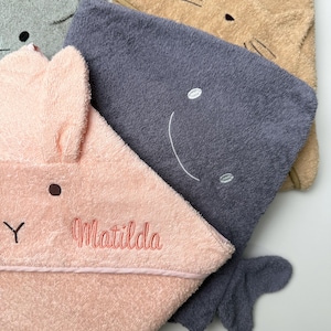 Op de afbeelding: Close-up van verschillende babyhanddoeken met capuchon in verschillende kleuren. Eén is roze met konijnenoren en de naam "Matilda" erop geborduurd. Een andere is grijs met een lachend gezicht. De handdoeken zijn zacht en pluizig.