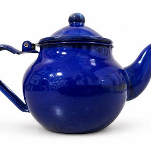 May include: A glossy, deep cobalt blue enamel teapot with a rounded body, a curved spout, and a matching lid featuring a small knob handle. The teapot has a sturdy, hinged metal handle attached to the side, finished in a matching blue enamel coating.