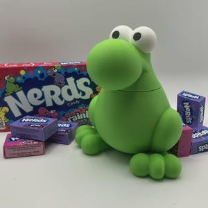 May include: A green frog-shaped container with large white eyes and a box of Nerds candy. Several boxes of Nerds candy in various flavors are scattered around the frog container. The box of candy has the word "Nerds" in a colorful design.