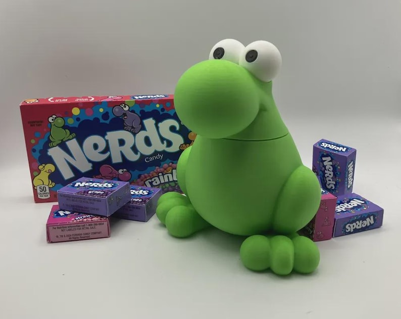May include: A green frog-shaped container with large white eyes and a box of Nerds candy. Several boxes of Nerds candy in various flavors are scattered around the frog container. The box of candy has the word "Nerds" in a colorful design.