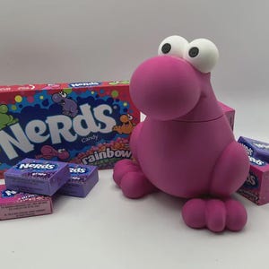 May include: A pink, cartoonish creature with large white eyes and a round snout, surrounded by boxes of Nerds candy. The candy boxes are various colors, including purple and pink, with the Nerds logo prominently displayed.