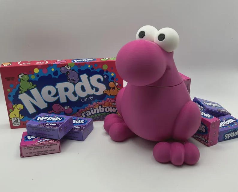 May include: A pink, cartoonish creature with large white eyes and a round snout, surrounded by boxes of Nerds candy. The candy boxes are various colors, including purple and pink, with the Nerds logo prominently displayed.
