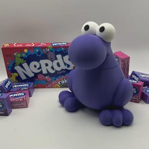 May include: A purple, cartoon-like creature with large white eyes and a round body, surrounded by boxes of Nerds candy. The candy boxes are various colors, including pink and purple, with the Nerds logo prominently displayed.