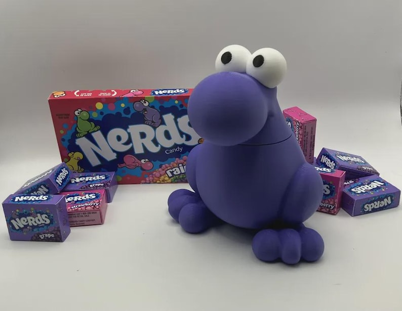 May include: A purple, cartoon-like creature with large white eyes and a round body, surrounded by boxes of Nerds candy. The candy boxes are various colors, including pink and purple, with the Nerds logo prominently displayed.