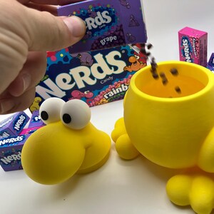 May include: A yellow, cartoon-like container with large white eyes and a round body, designed to resemble a character. Boxes of Nerds candy in various flavors, including grape and strawberry, are visible. Candy is being poured into the container.