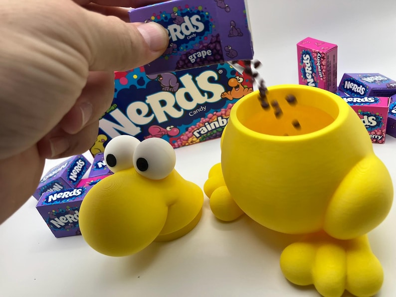 May include: A yellow, cartoon-like container with large white eyes and a round body, designed to resemble a character. Boxes of Nerds candy in various flavors, including grape and strawberry, are visible. Candy is being poured into the container.