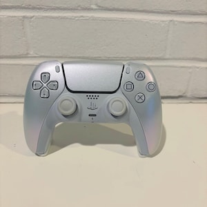 Pode incluir: Um comando sem fios DualSense branco para PlayStation 5 com acabamento perolado. O comando tem um logotipo PlayStation preto e um touchpad preto.