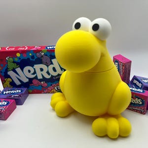 May include: A bright yellow, cartoonish creature-shaped container with large white eyes and a round head. The container is surrounded by boxes of Nerds candy in various flavors, including grape and strawberry. A large box of Nerds candy is in the background.