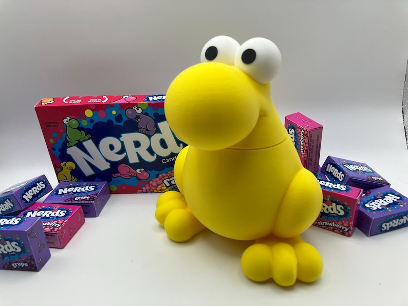 May include: A bright yellow, cartoonish creature-shaped container with large white eyes and a round head. The container is surrounded by boxes of Nerds candy in various flavors, including grape and strawberry. A large box of Nerds candy is in the background.