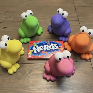 May include: Five colorful frog-like figurines with large, white-eyed features are arranged around a box of "Nerds" candy. The figurines are in shades of green, purple, orange, yellow, and pink. The candy box has a blue and red design.