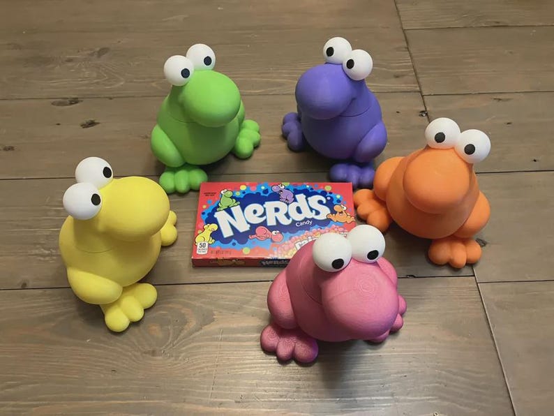 May include: Five colorful frog-like figurines with large, white-eyed features are arranged around a box of "Nerds" candy. The figurines are in shades of green, purple, orange, yellow, and pink. The candy box has a blue and red design.