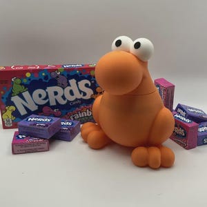 May include: An orange, cartoon-like figure with large white eyes and small feet and hands. The figure is surrounded by boxes of Nerds candy, including a large box and several smaller boxes of various flavors. The large box reads "Nerds Candy".