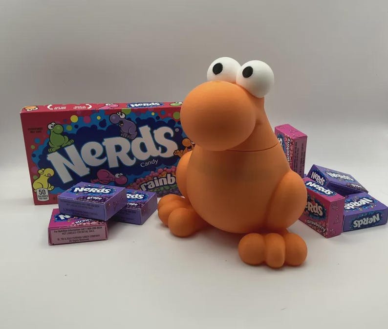 May include: An orange, cartoon-like figure with large white eyes and small feet and hands. The figure is surrounded by boxes of Nerds candy, including a large box and several smaller boxes of various flavors. The large box reads "Nerds Candy".