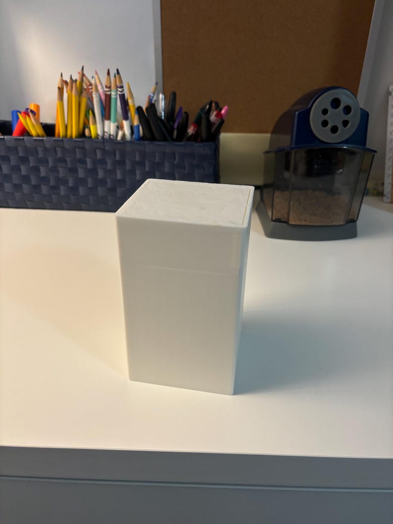 May include: A white, square-shaped box with a smooth, matte finish. The box is sitting on a white surface.