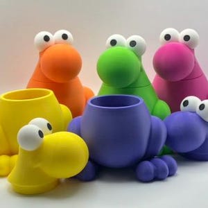 May include: A collection of six whimsical, colorful creature-shaped containers. The containers are in shades of yellow, orange, green, pink, purple, and blue. Each has large, white-eyed, round faces and a cylindrical opening.