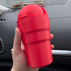 May include: A bright red, cylindrical car trash can with a hinged lid. The trash can has a ridged design and is held in a hand. The lid has two small, circular features. The background is a car interior.