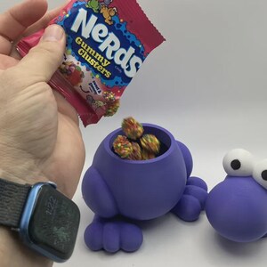 May include: A purple, frog-shaped container with large white eyes, filled with colorful Nerds Gummy Clusters candy. A hand holds a bag of Nerds Gummy Clusters, pouring candy into the container. The bag has the text "Nerds Gummy Clusters".