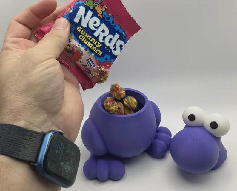 May include: A purple, frog-shaped container with large white eyes, filled with colorful Nerds Gummy Clusters candy. A hand holds a bag of Nerds Gummy Clusters, pouring candy into the container. The bag has the text "Nerds Gummy Clusters".