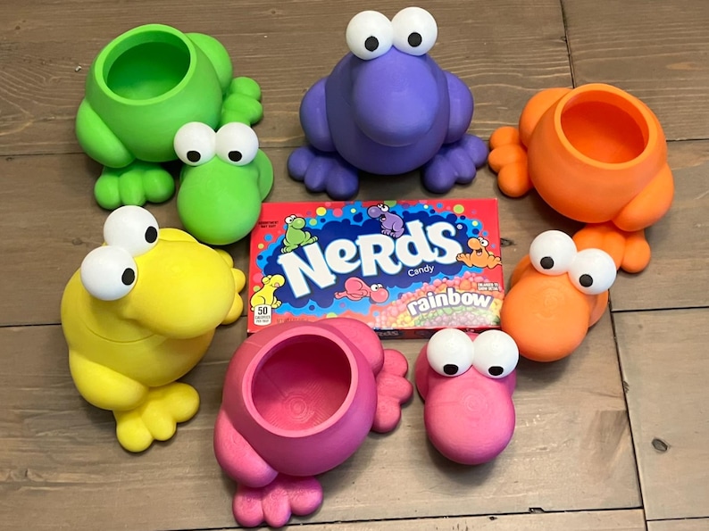 May include: A collection of colorful, frog-shaped containers in green, purple, orange, yellow, and pink, each with large, googly eyes. A box of Nerds Rainbow candy is in the center.